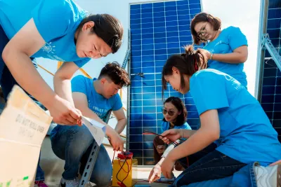 Young participants wearing blue shirts work together outdoors to assemble and test solar power equipment. They are gathered around wiring and tools beside large solar panels, concentrating closely on the task. A UNICEF Mongolia logo appears in the corner.