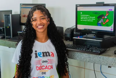 A smiling young woman with long curly hair stands in a computer lab, wearing a white IEMA T-shirt. Behind her, desktop computers display educational content on their screens.