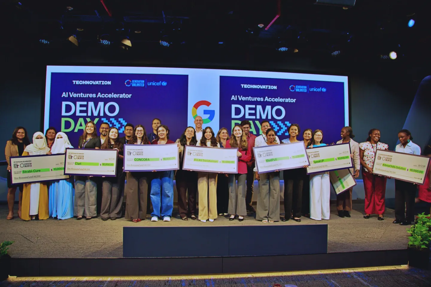 AIVA trainees, young women from diverse countries, stand on stage holding placards with their funding money in front of an audience at Demo Day in India