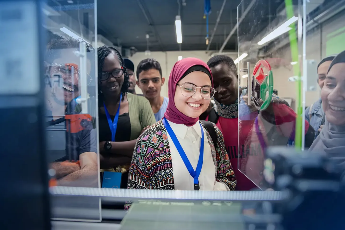 A group of young adults wearing ID lanyards stand closely together in a modern lab or workshop space, looking through a glass enclosure at equipment. A smiling woman in a pink headscarf and glasses stands at the center, while others around her focus on the same project.
