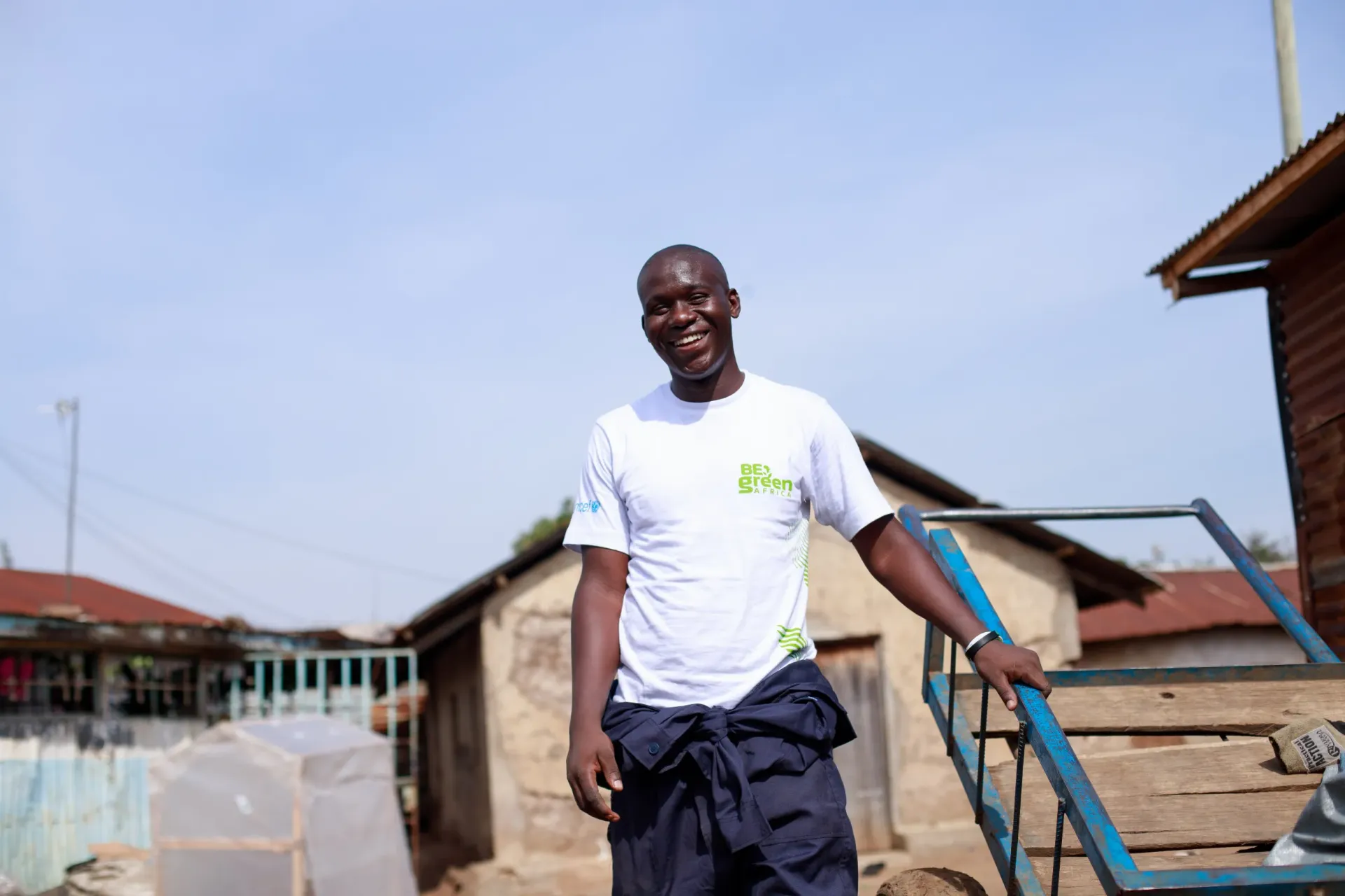 Meshack Ogutu stands outdoors in a modest residential area with simple buildings and tin-roofed structures in the background.