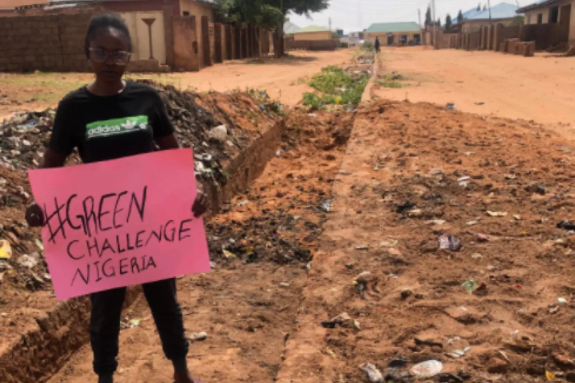 A young Nigerian girl stands on a dirt road holding a sign about the green challenge