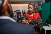 woman wearing a colorful patterned outfit and headwrap gestures while speaking to attendees in a meeting room.