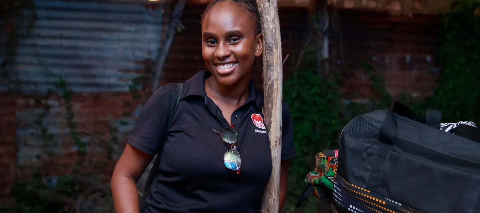 A woman smiles at the camera while standing beside a table displaying handmade bags in an outdoor workspace.
