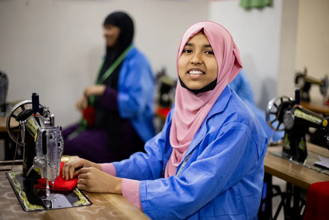 A young woman wearing a pink headscarf smiles while operating a sewing machine during a skills training session in Bangladesh