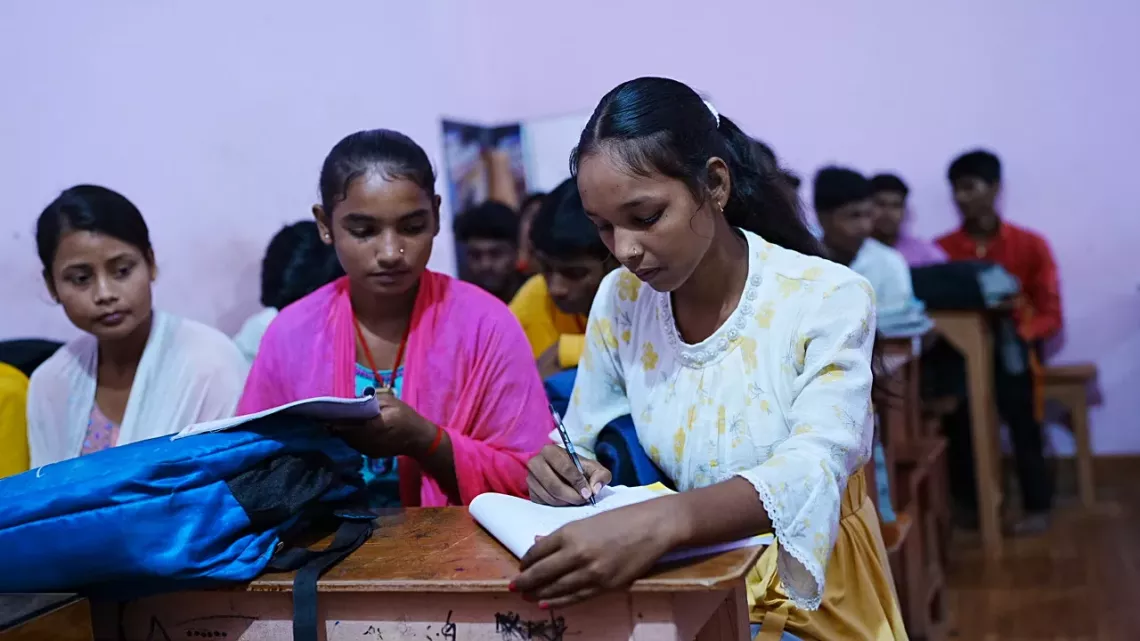Muskaan, aged 15, in a classroom surrounded by fellow students. She is reading her notebook alongside a classmate, engaged in the learning environment.