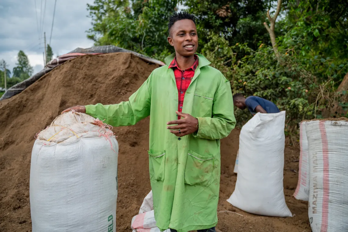 Kennedy shown in a green farming trenchcoat with sacks of either coffe, beans, avocado or tomatoes