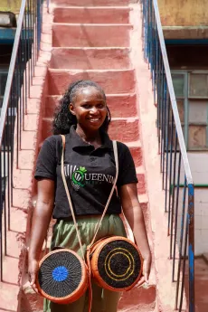 A smiling young woman standing on outdoor steps, holding handcrafted artisanal products.