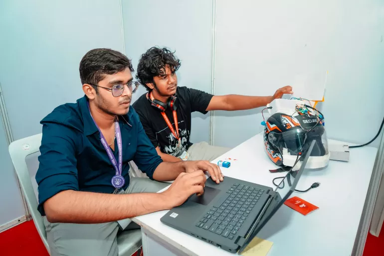 Two adolescents fromIndia working at a table. One is focused on a laptop, while the other adjusts a helmet with electronic components and wires attached.