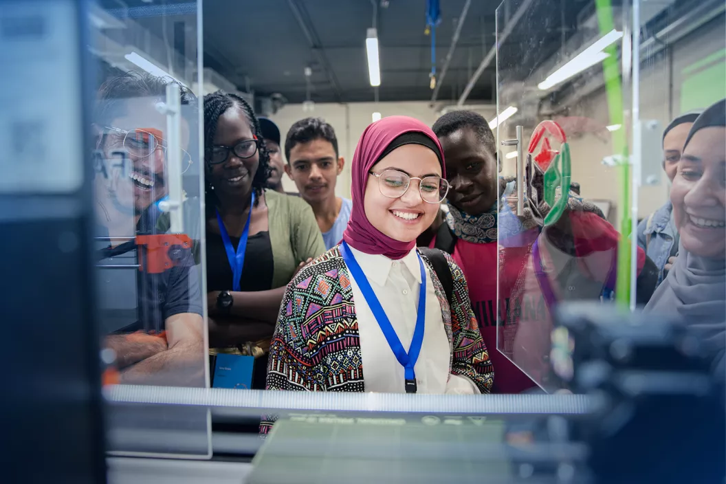 At the imagen ventures global workshop in Jordan, a young girl is smiling looking down at a 3D printer, with a number of friends and colleageus behind her 