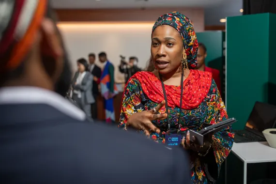 woman wearing a colorful patterned outfit and headwrap gestures while speaking to attendees in a meeting room.