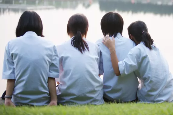 Four girls sit together on grass, with their backs to the camera, as they look out over water.